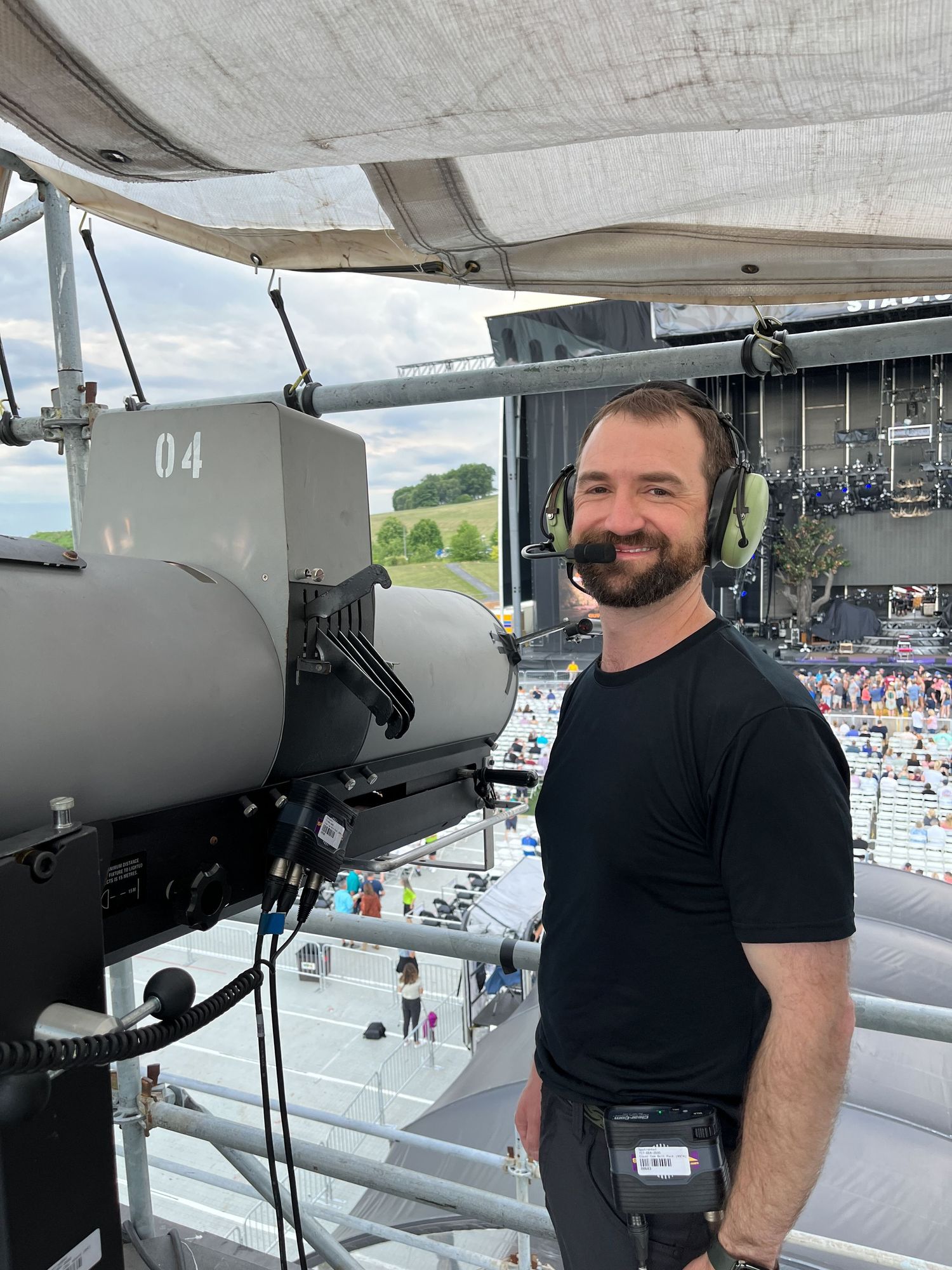 Man in black shirt smiles while wearing a headset, standing by a spotlight tower at an outdoor concert venue.