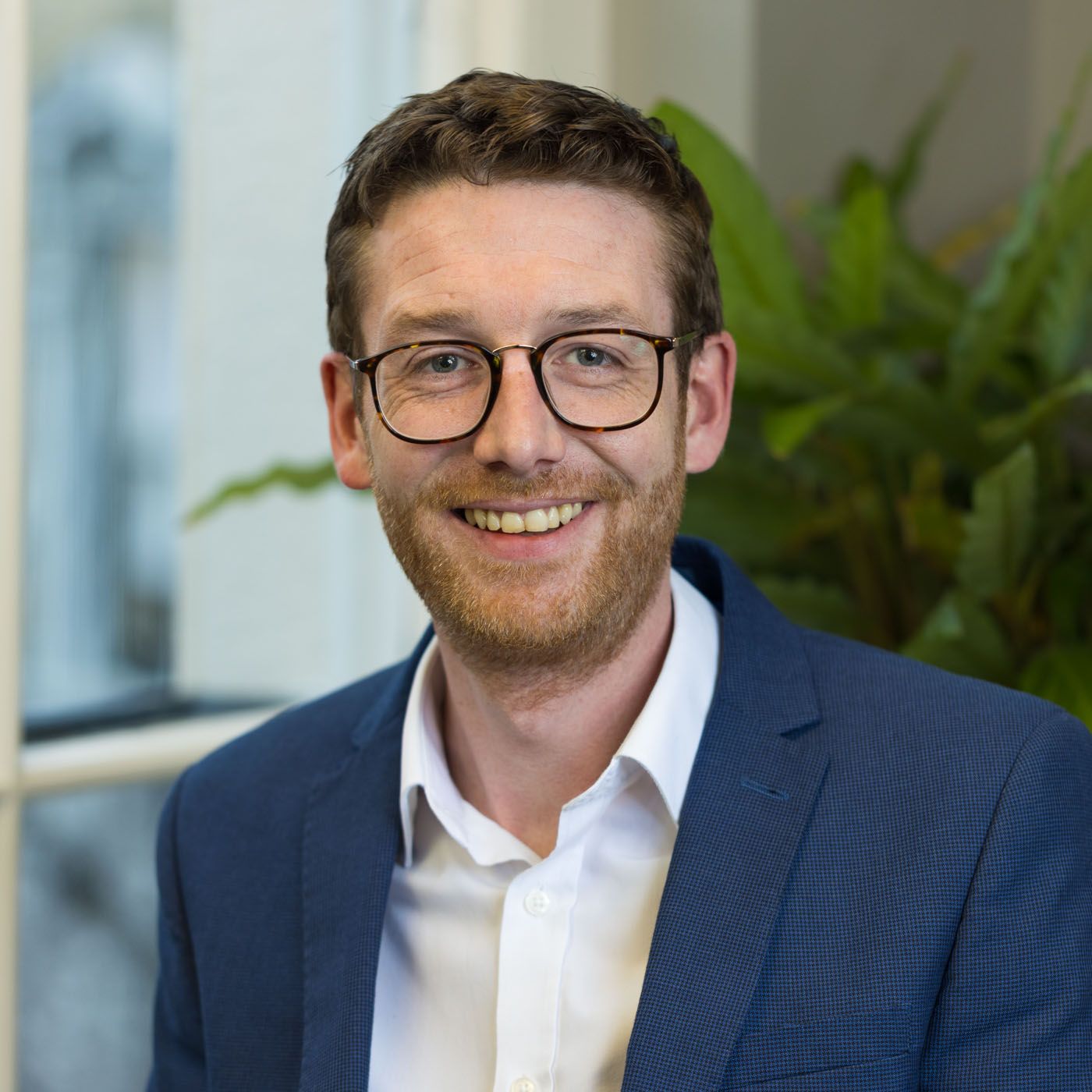 Professional headshot of someone in glasses and navy suit with white shirt smiling warmly with green plant in background.