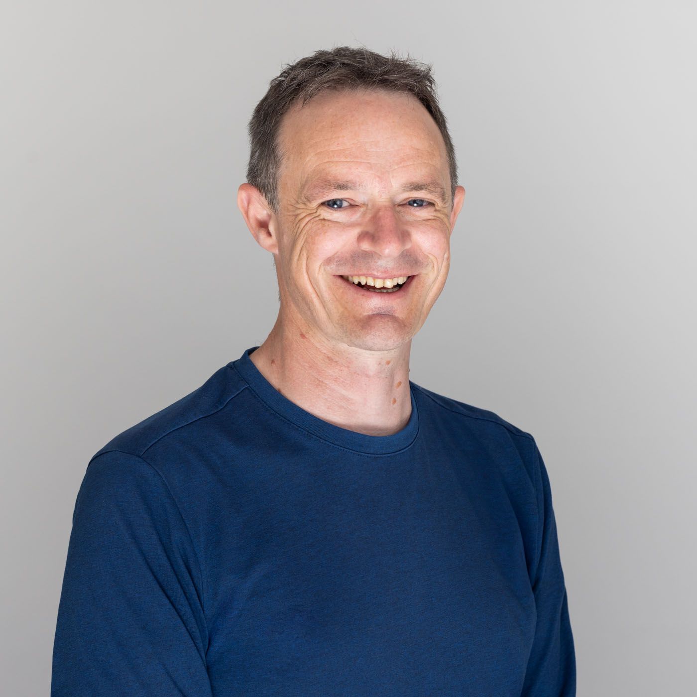 A person wearing a navy blue t-shirt smiles warmly against a light gray background in a professional headshot.