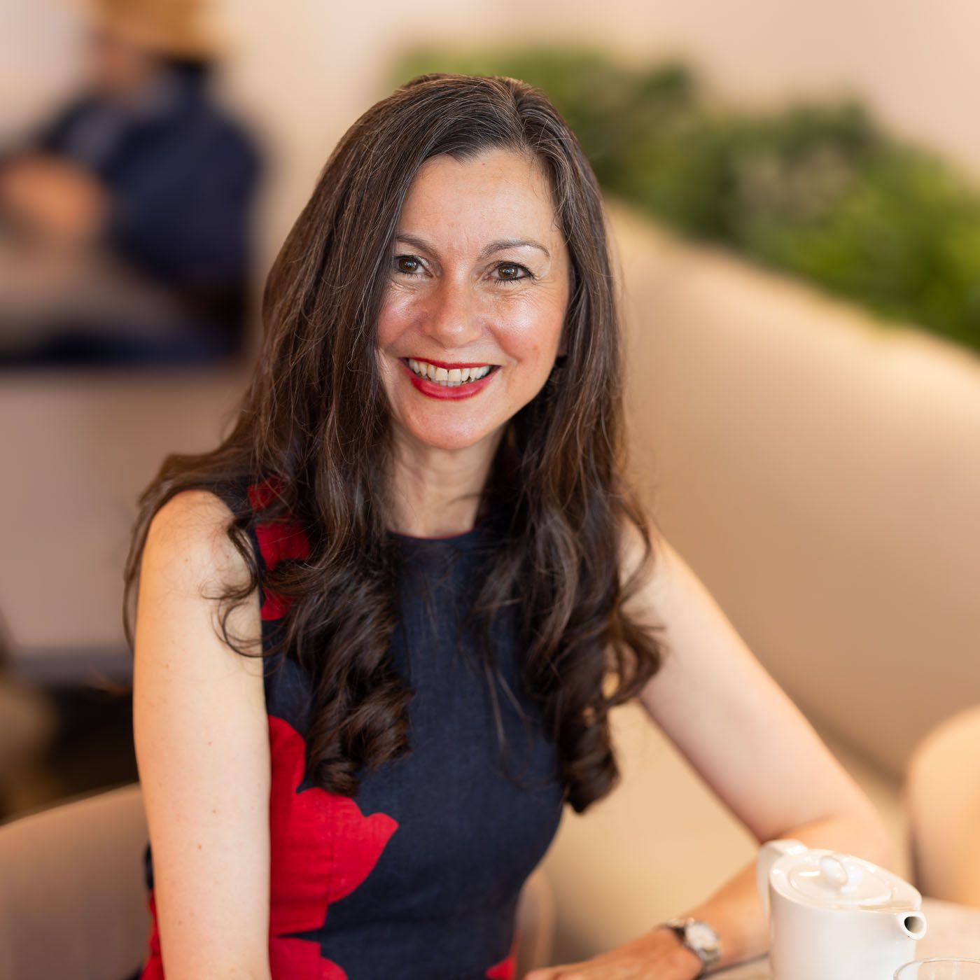 Professional portrait of person in navy and red dress sitting at table with coffee, smiling warmly.