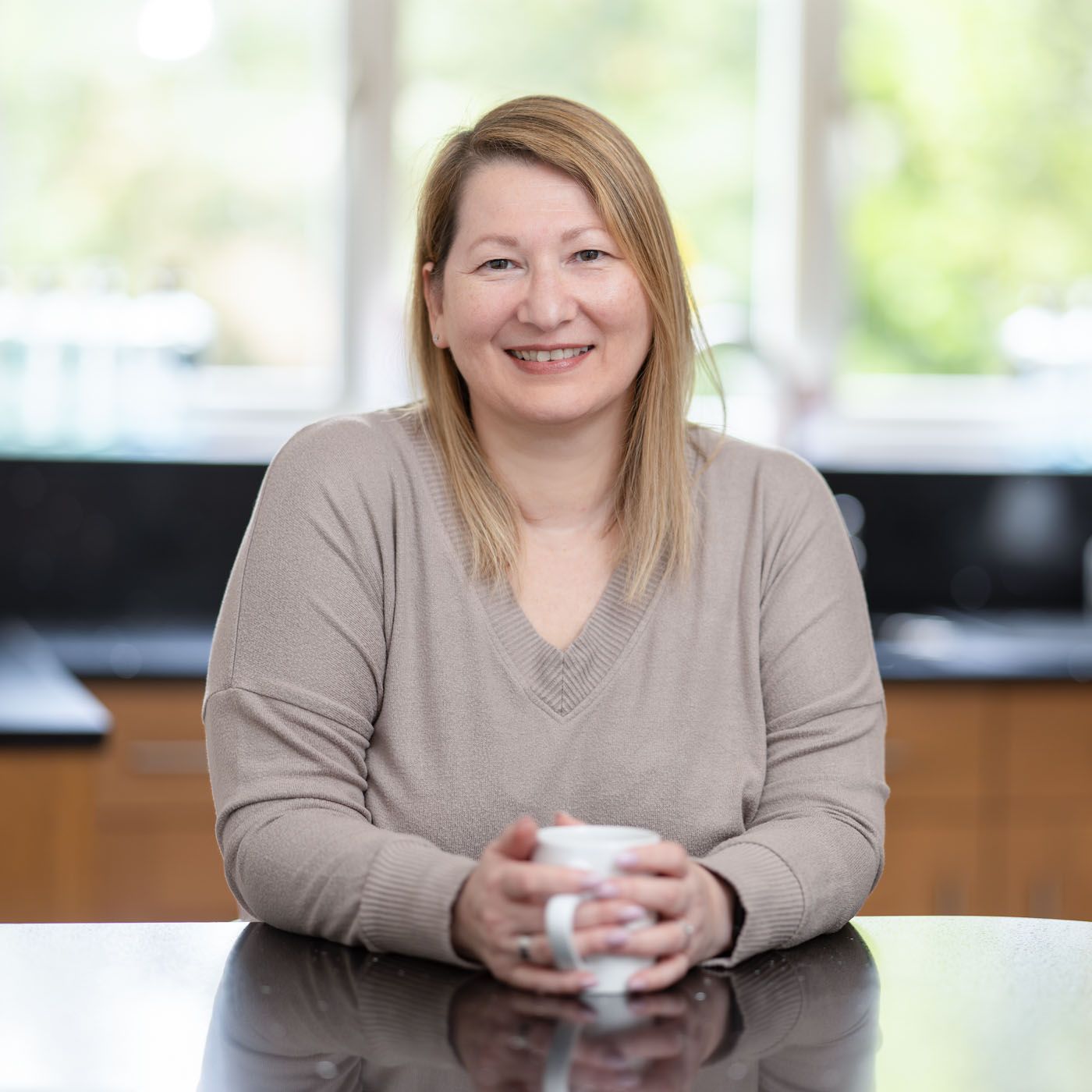 Someone sits at a desk holding a white coffee mug while wearing a beige v-neck sweater and smiling warmly at the camera.