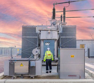 Industrial and engineering photographer | ABB engineer in high-visibility vest inspecting electrical transformer substation with sunset sky.