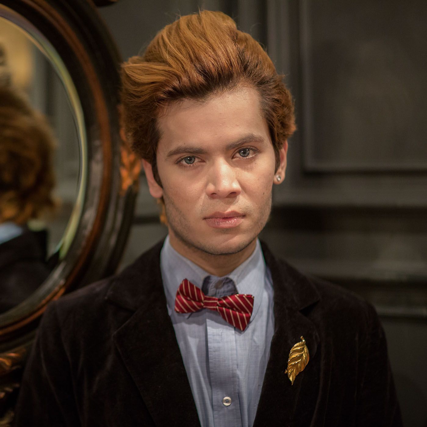 A well-dressed person with styled reddish hair wearing a velvet blazer, striped bow tie and gold brooch.