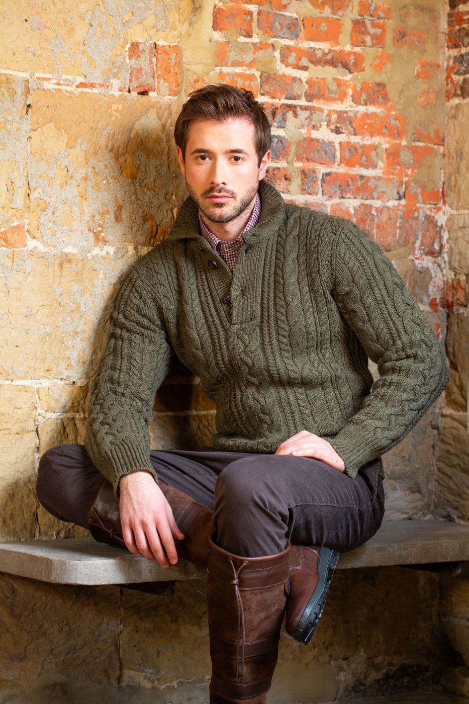 Fashionable cable knit sweater in olive green styled with brown boots against rustic brick wall background.