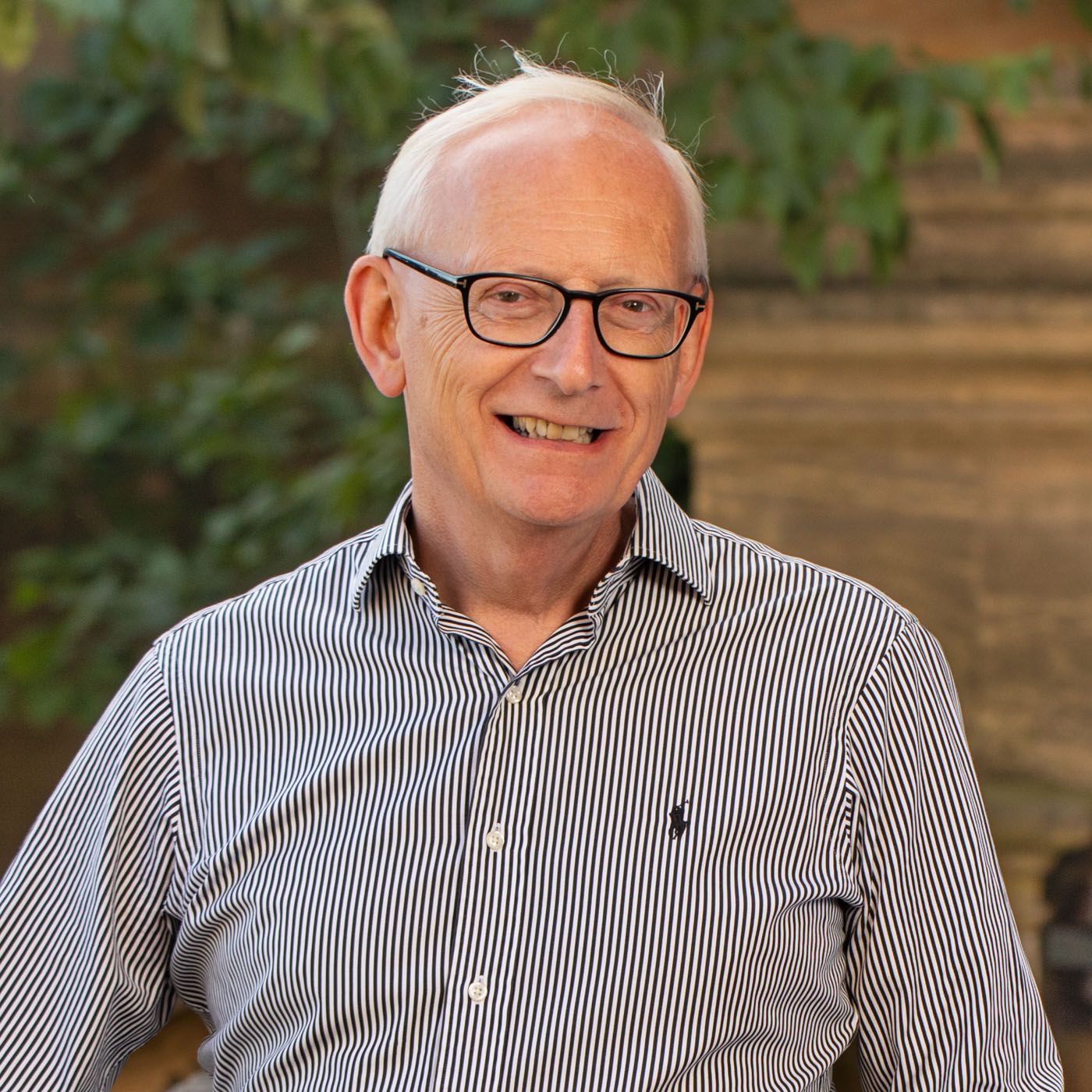 Smiling senior wearing glasses and striped button-down shirt poses outdoors with natural backdrop.