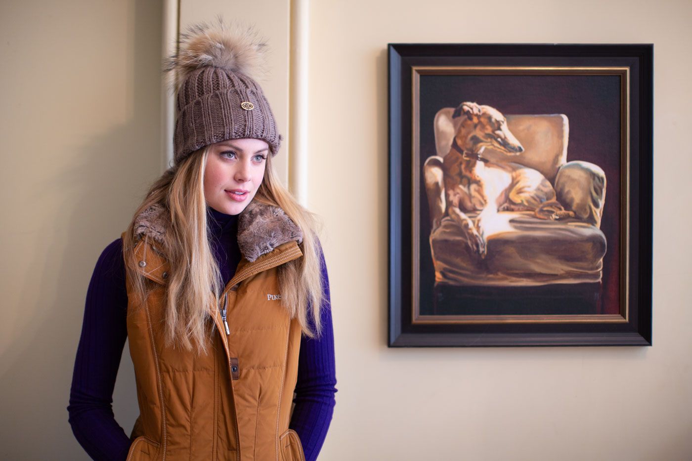 Someone wearing a brown vest and knit beanie stands against a wall with framed artwork hanging in the background.