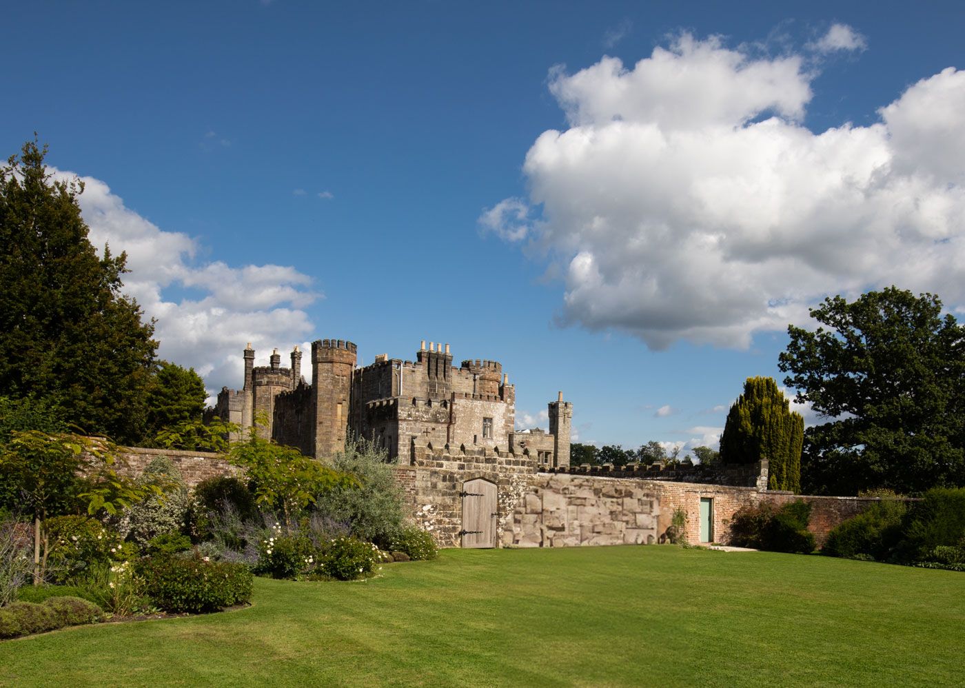 Medieval stone castle with turrets and towers sits behind manicured gardens on a sunny day with white puffy clouds.