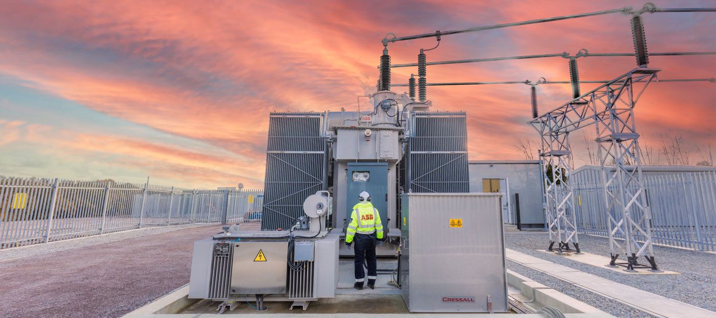 Maintenance worker inspects electrical substation equipment against dramatic orange sunset sky with power lines.