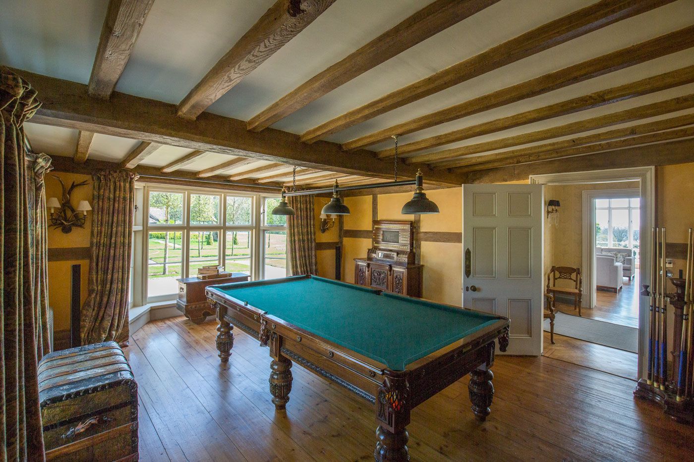 Elegant game room with a vintage pool table, exposed wooden beams, and large windows in a rustic country home setting.