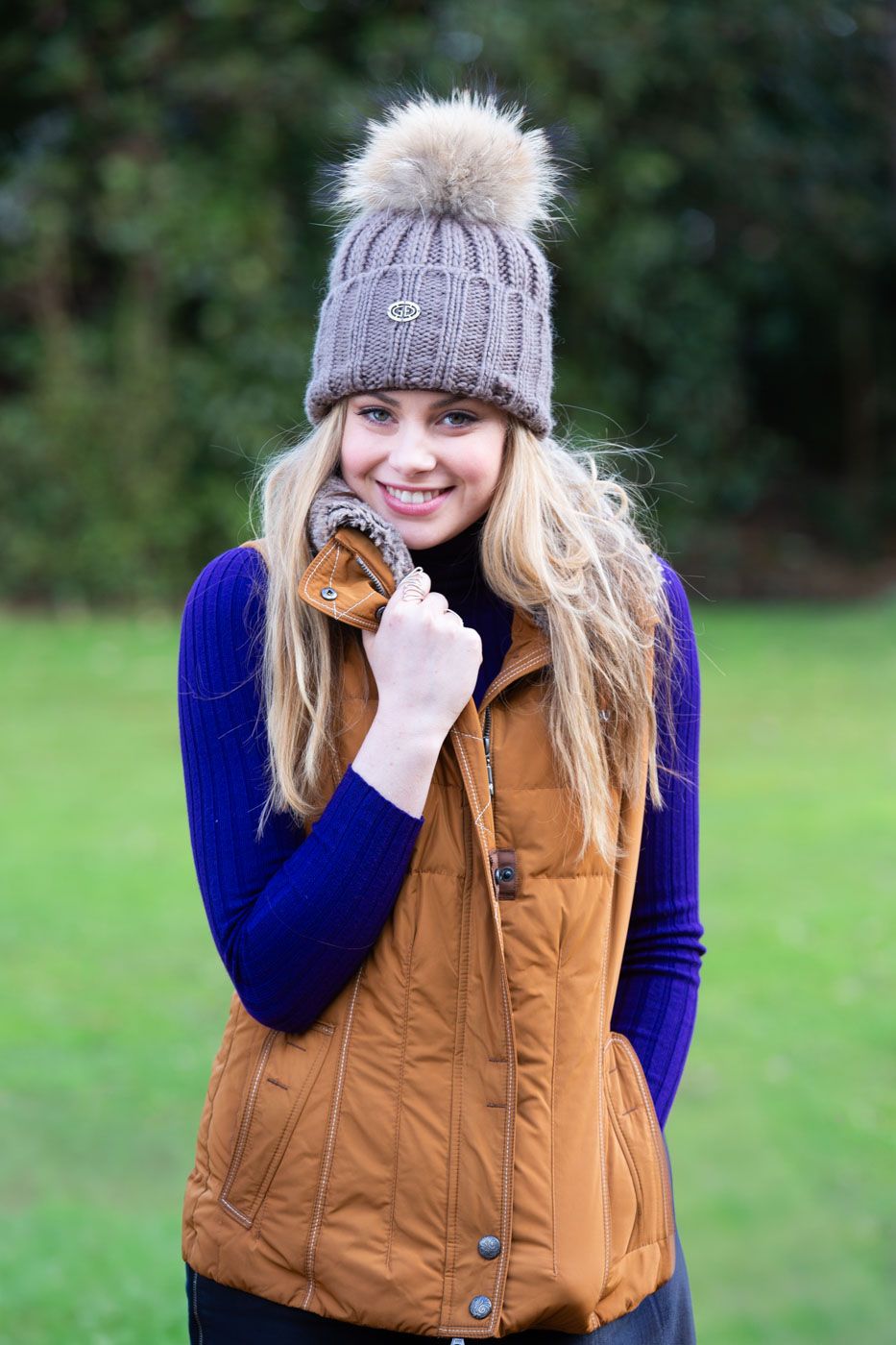 Someone wearing a gray knit pom beanie, blue sweater and brown puffy vest poses outdoors on a grassy field.