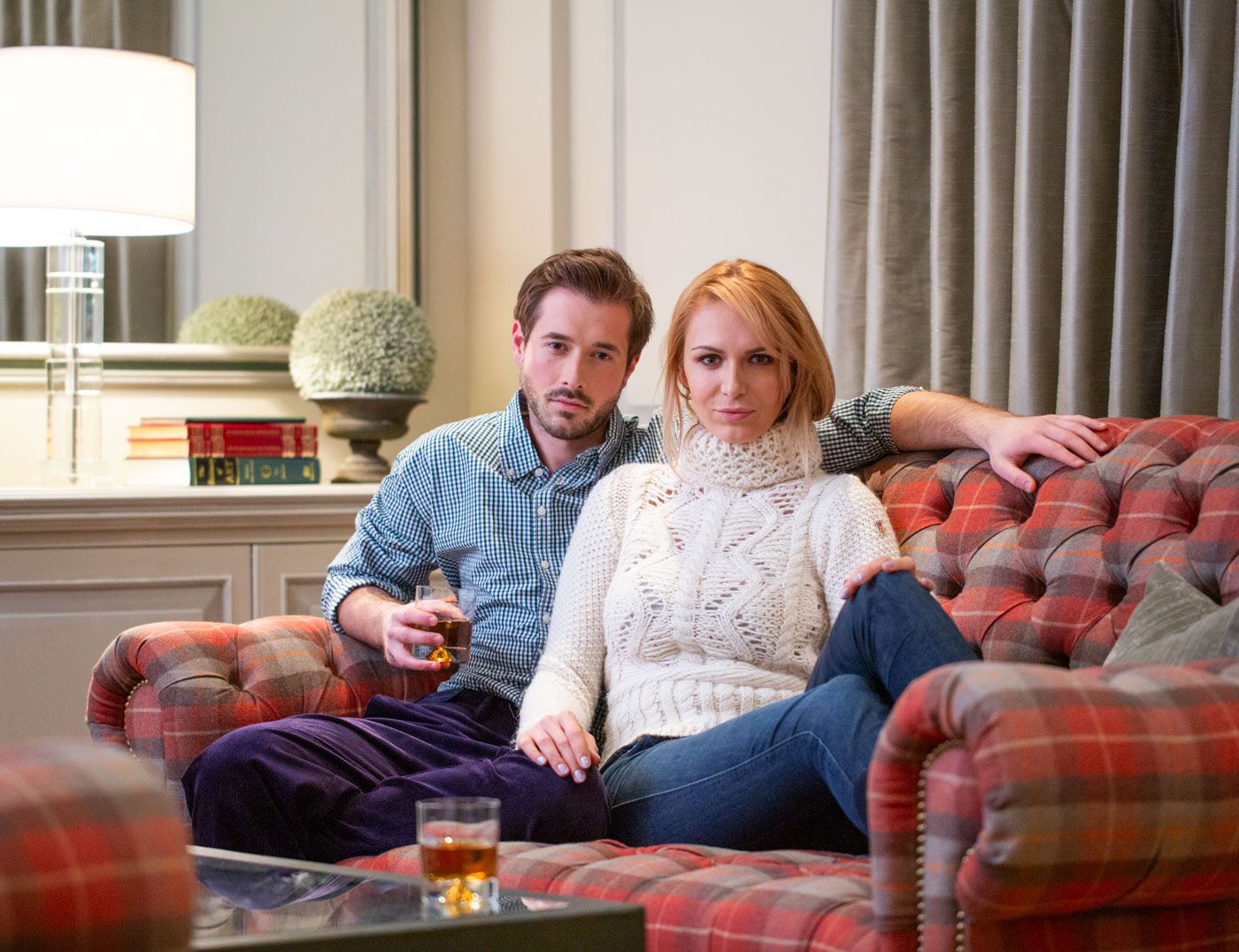 Two people relax together on a plush red plaid sofa with drinks nearby in a cozy living room setting.