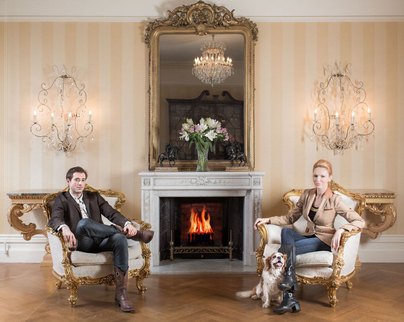 Elegant living room with ornate gold chairs, crystal sconces, marble fireplace and dog sitting on hardwood floors.