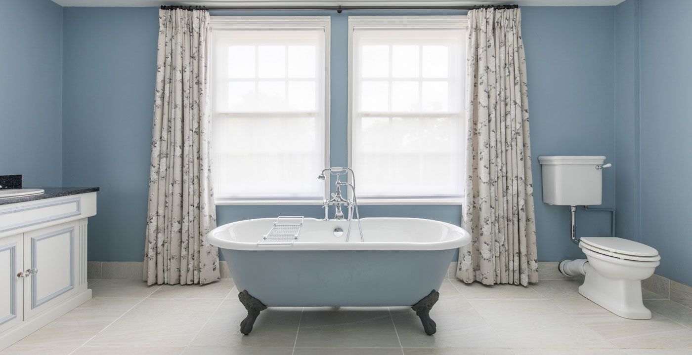 Elegant clawfoot bathtub in a light blue bathroom with floral curtains, white fixtures and tile flooring.