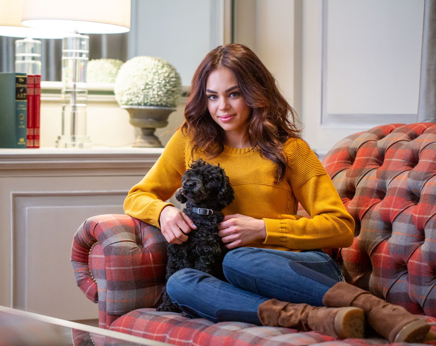 A person in a yellow sweater sits on a plush red couch while holding a small black dog.