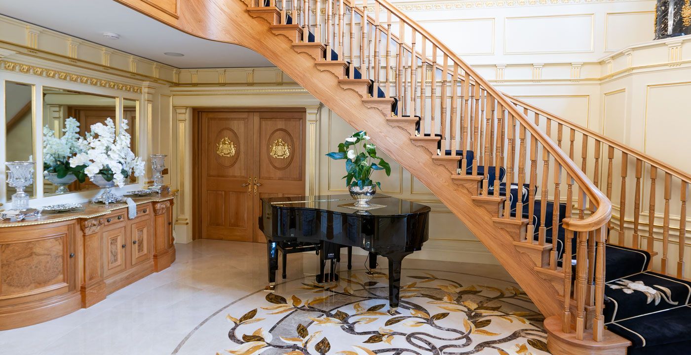 A grand foyer with a baby grand piano beneath a wooden staircase features elegant decor and a patterned rug.