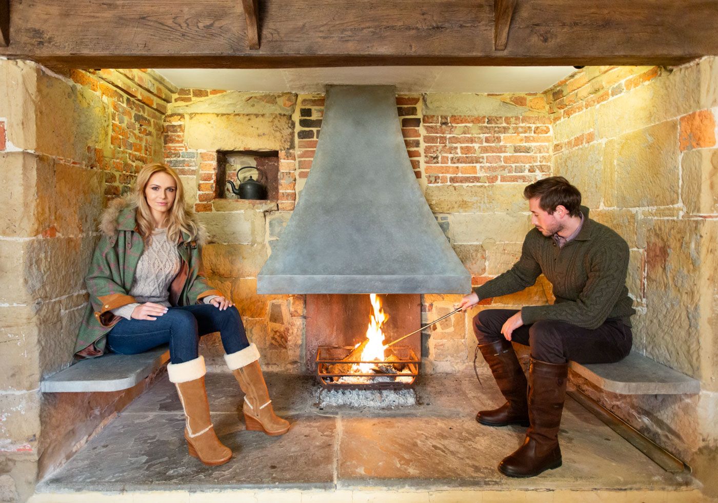 Two people sit in a cozy stone inglenook with exposed brick walls and a traditional fireplace between them, enjoying the warmth.