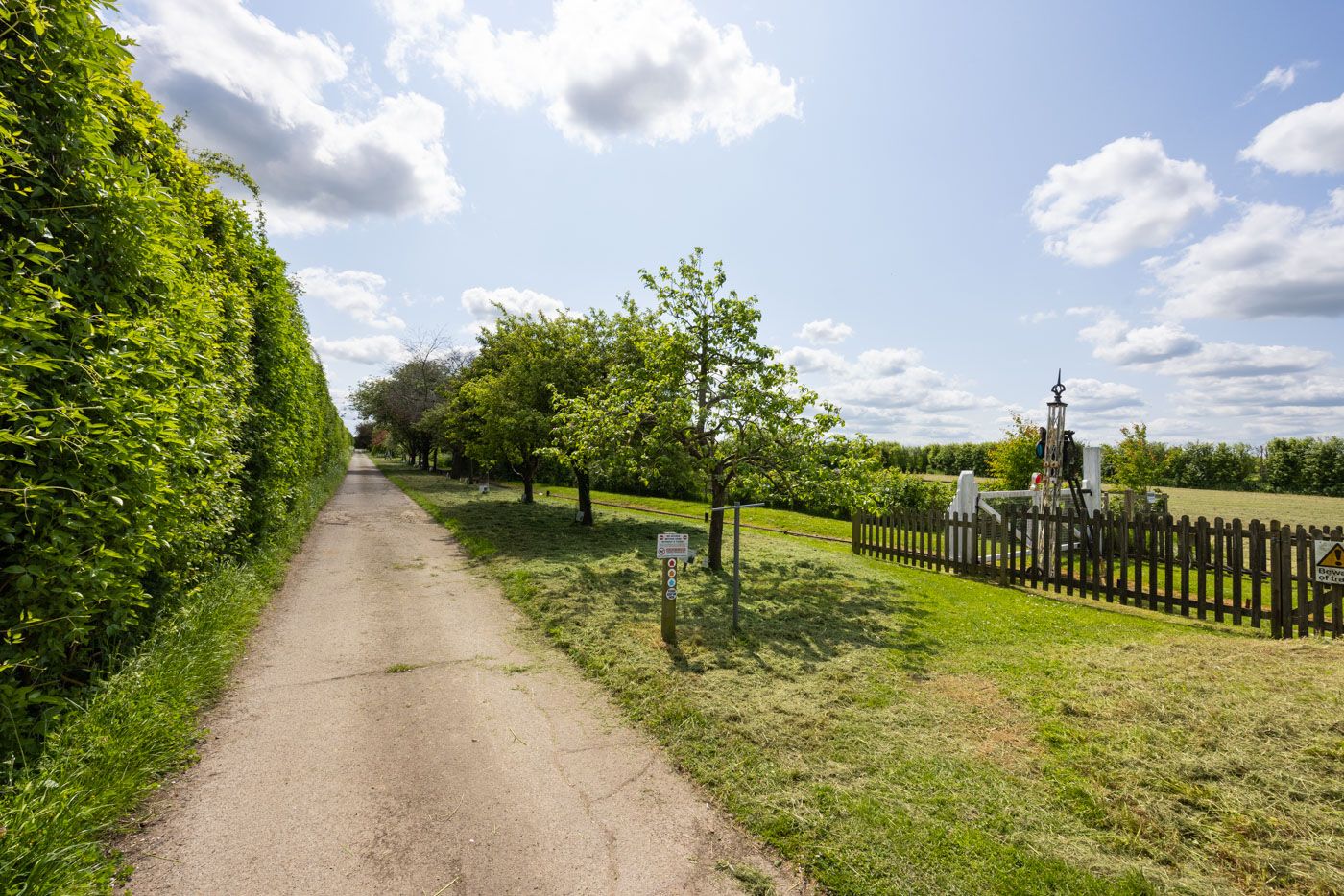 A winding countryside path lined with green trees and hedges leads past a wooden fence under a partly cloudy summer sky.