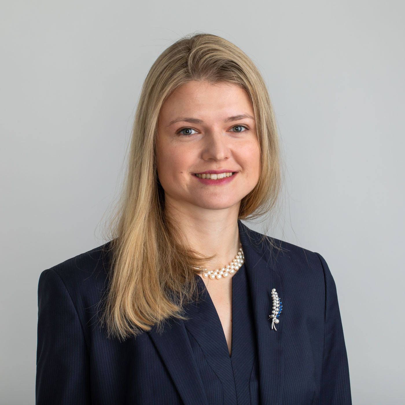 Professional headshot of a person in a navy blazer and pearl necklace smiling against a light gray background.