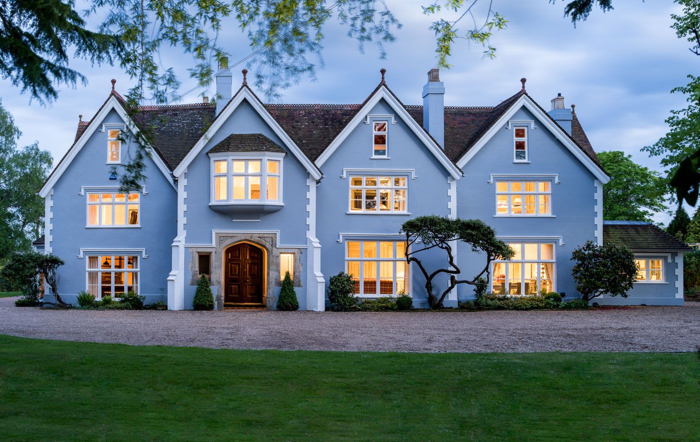 Large elegant blue Tudor-style mansion illuminated at dusk with multiple peaked gables and warm glowing windows.