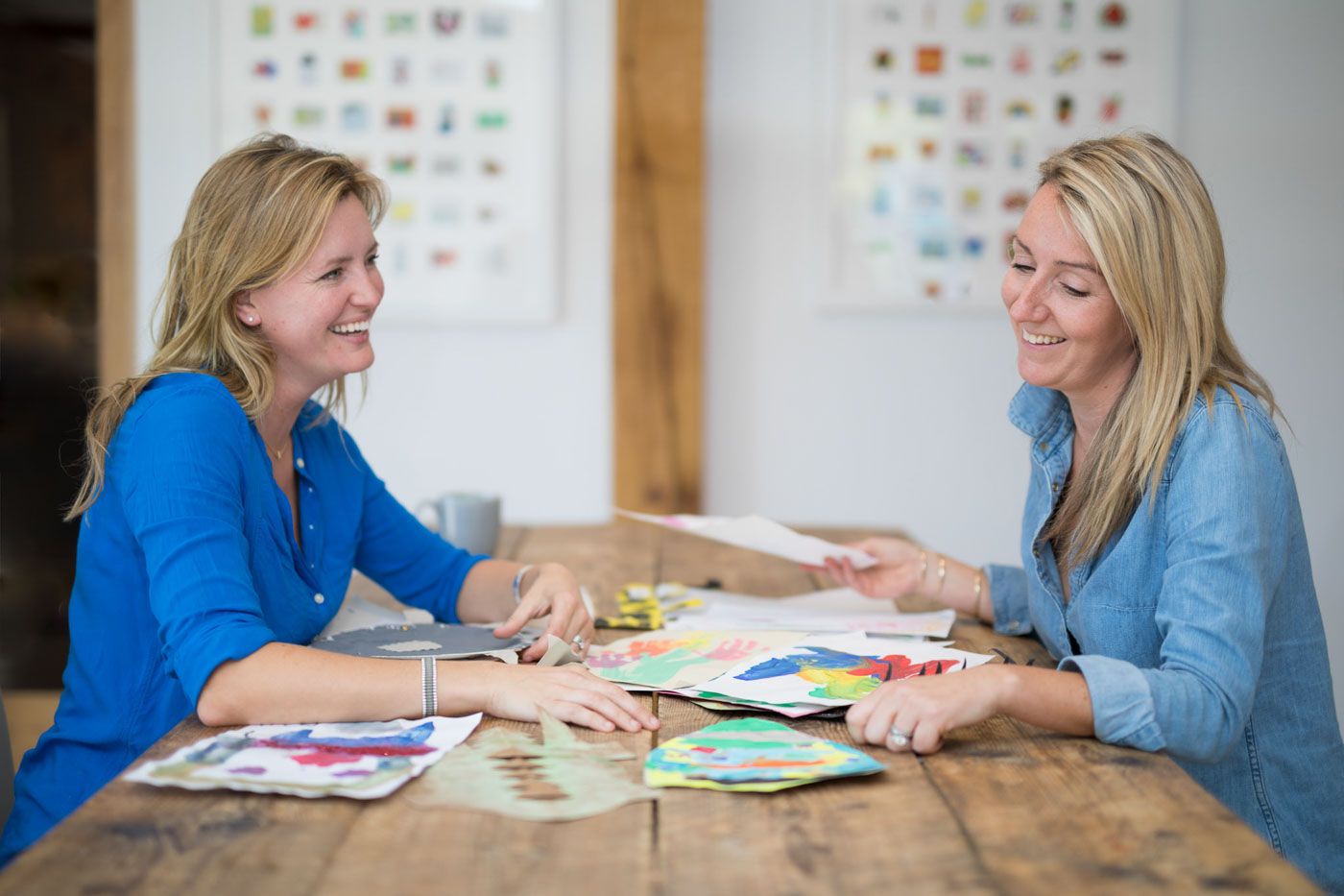 Two women in blue tops sit at wooden table working together with colourful papers and documents spread between them.