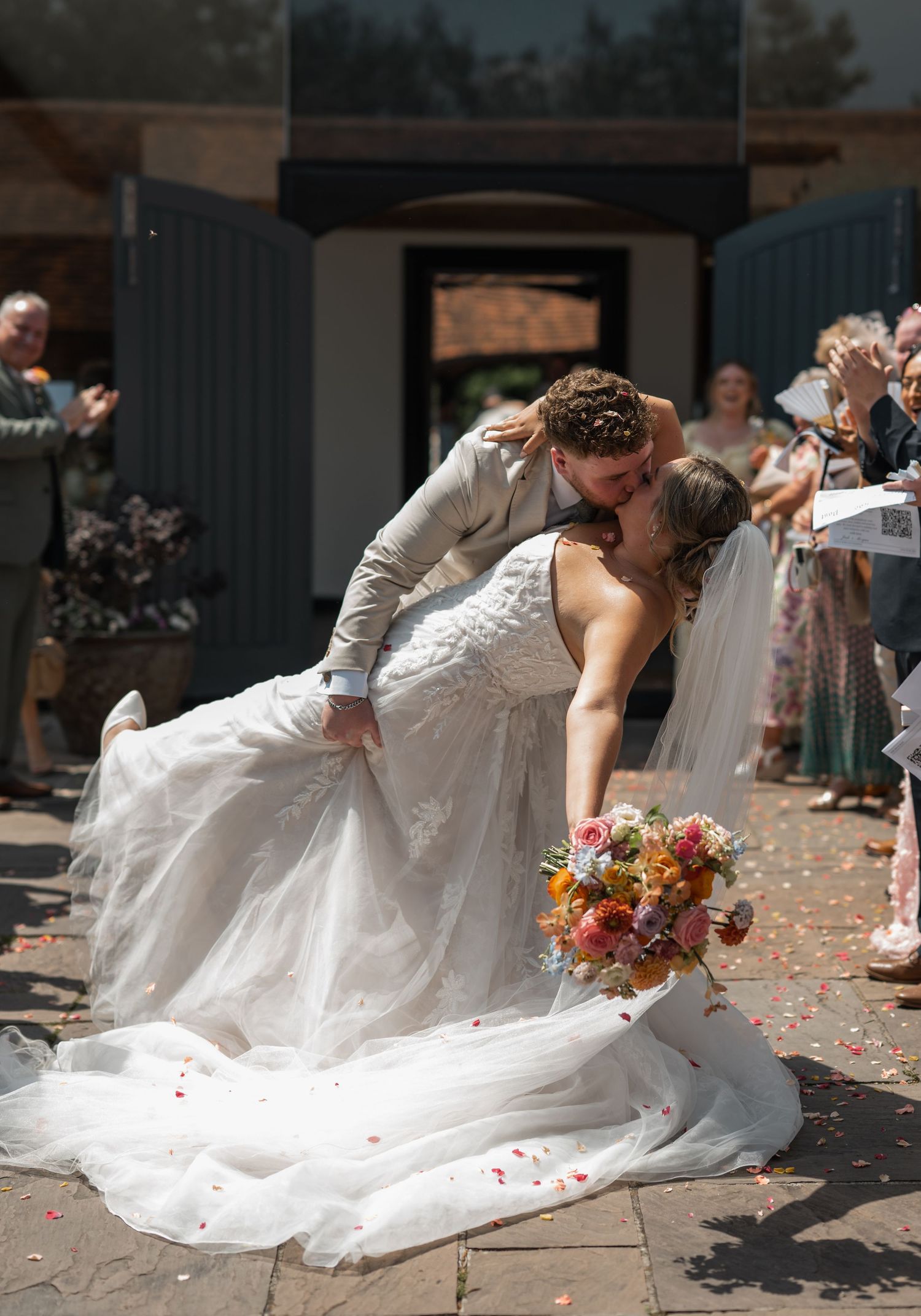 Bride and groom share a romantic dip kiss outside wedding venue as guests celebrate with colorful confetti.