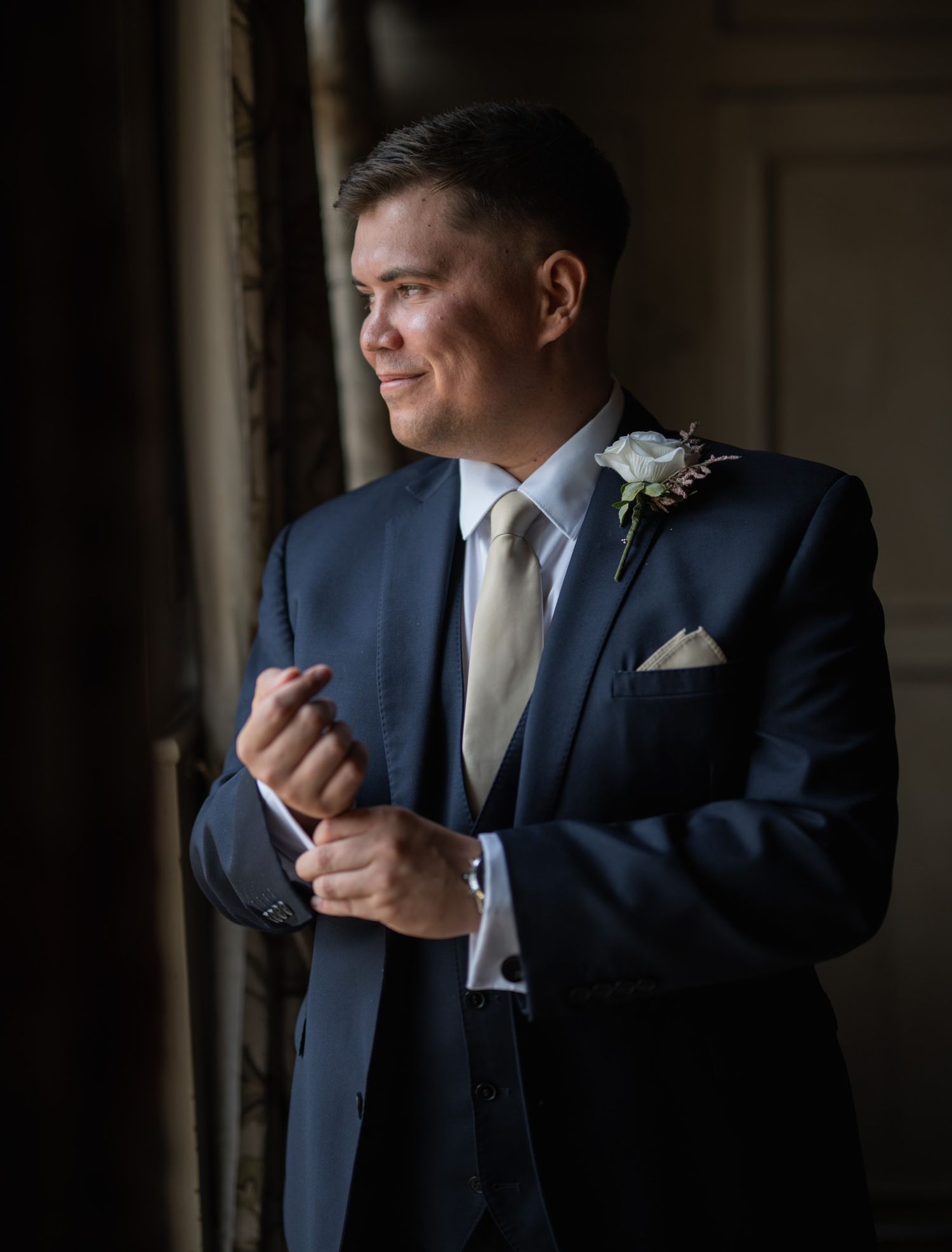 Groom in navy suit adjusting cufflinks by window, wearing white boutonniere and gold tie on wedding day.