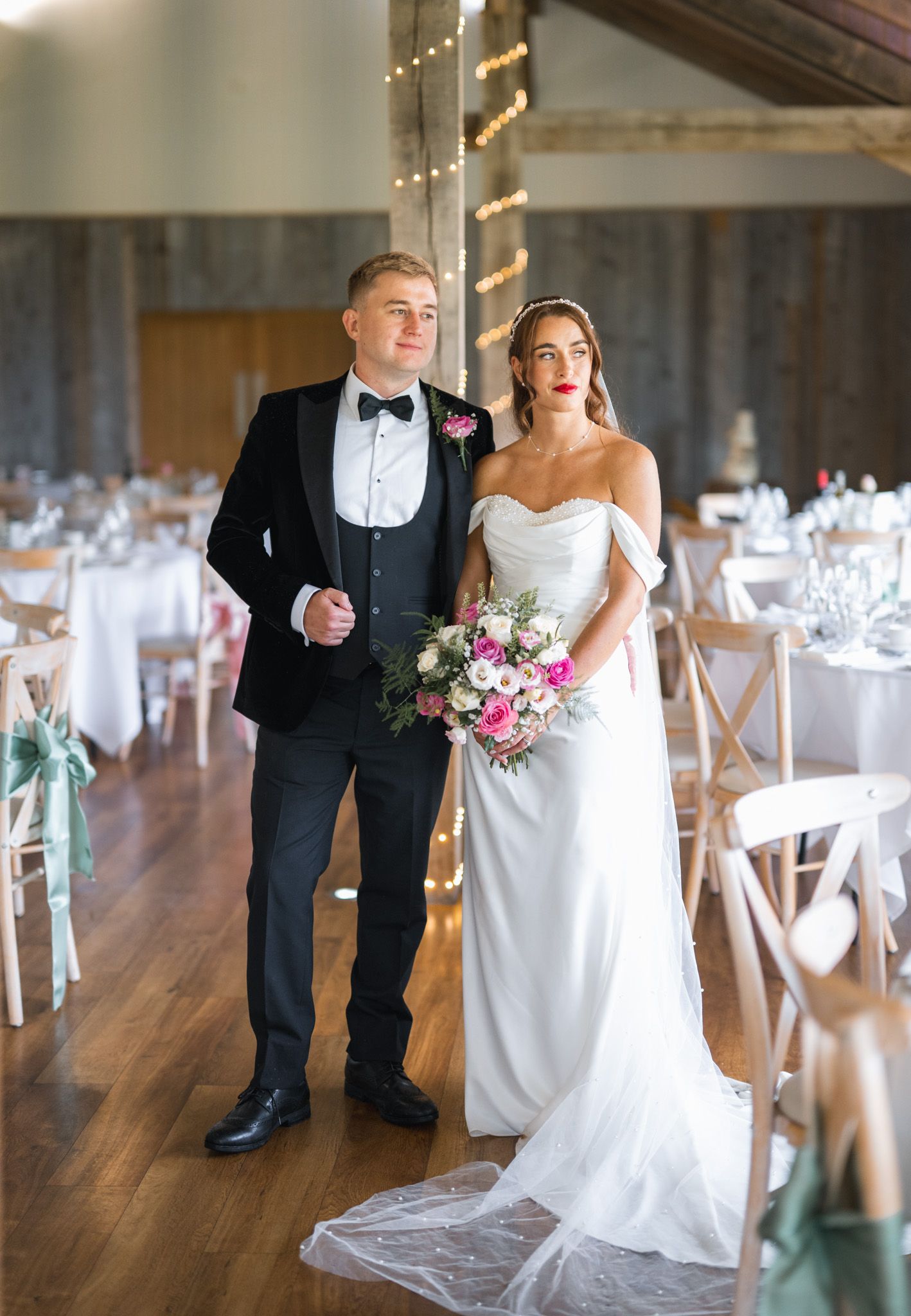 Bride in off-shoulder white gown holding pink bouquet stands with groom in black tuxedo in elegant barn reception venue.