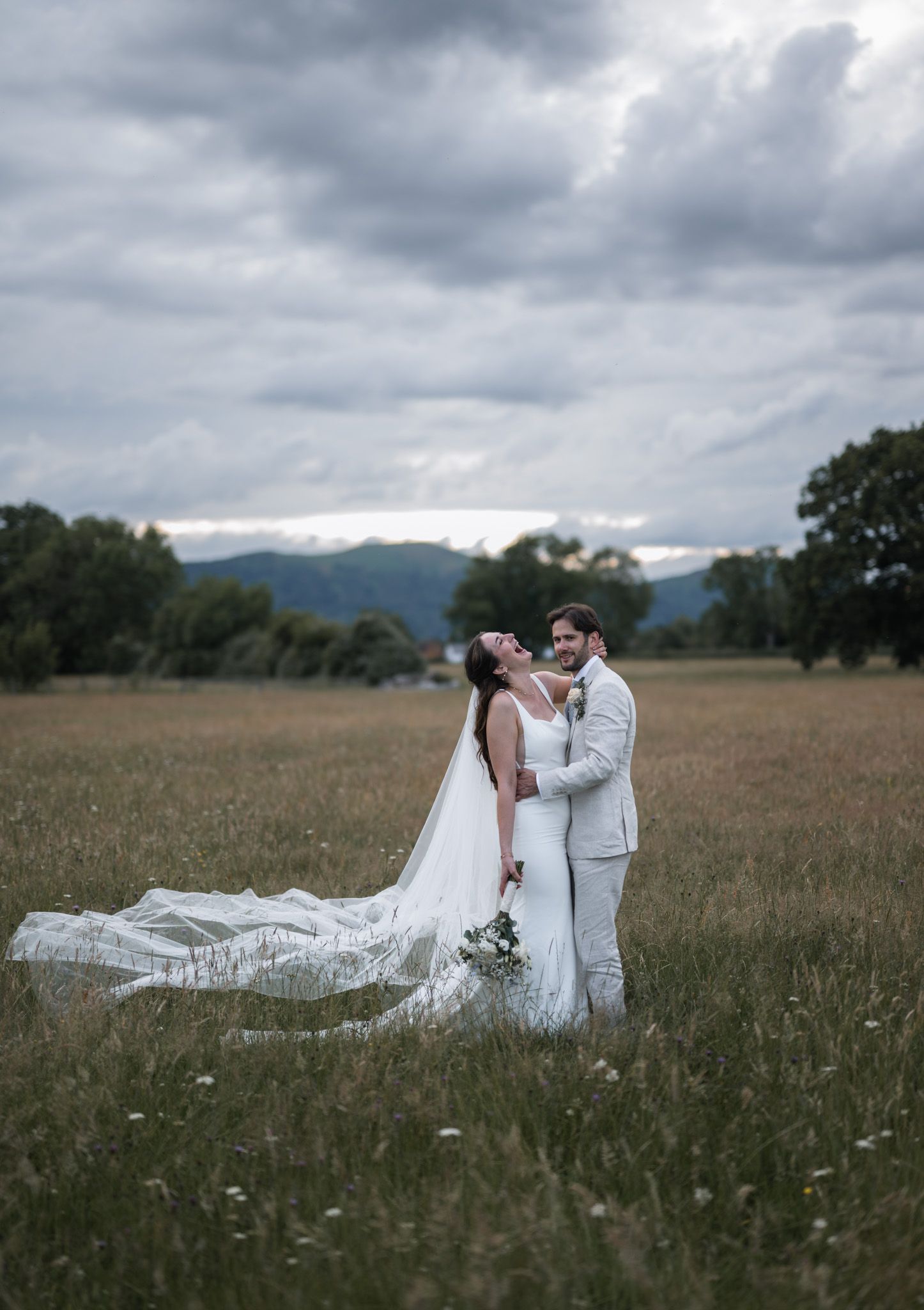 Bride and groom embrace in a scenic meadow, her veil flowing in the wind, with mountains and dramatic clouds behind.