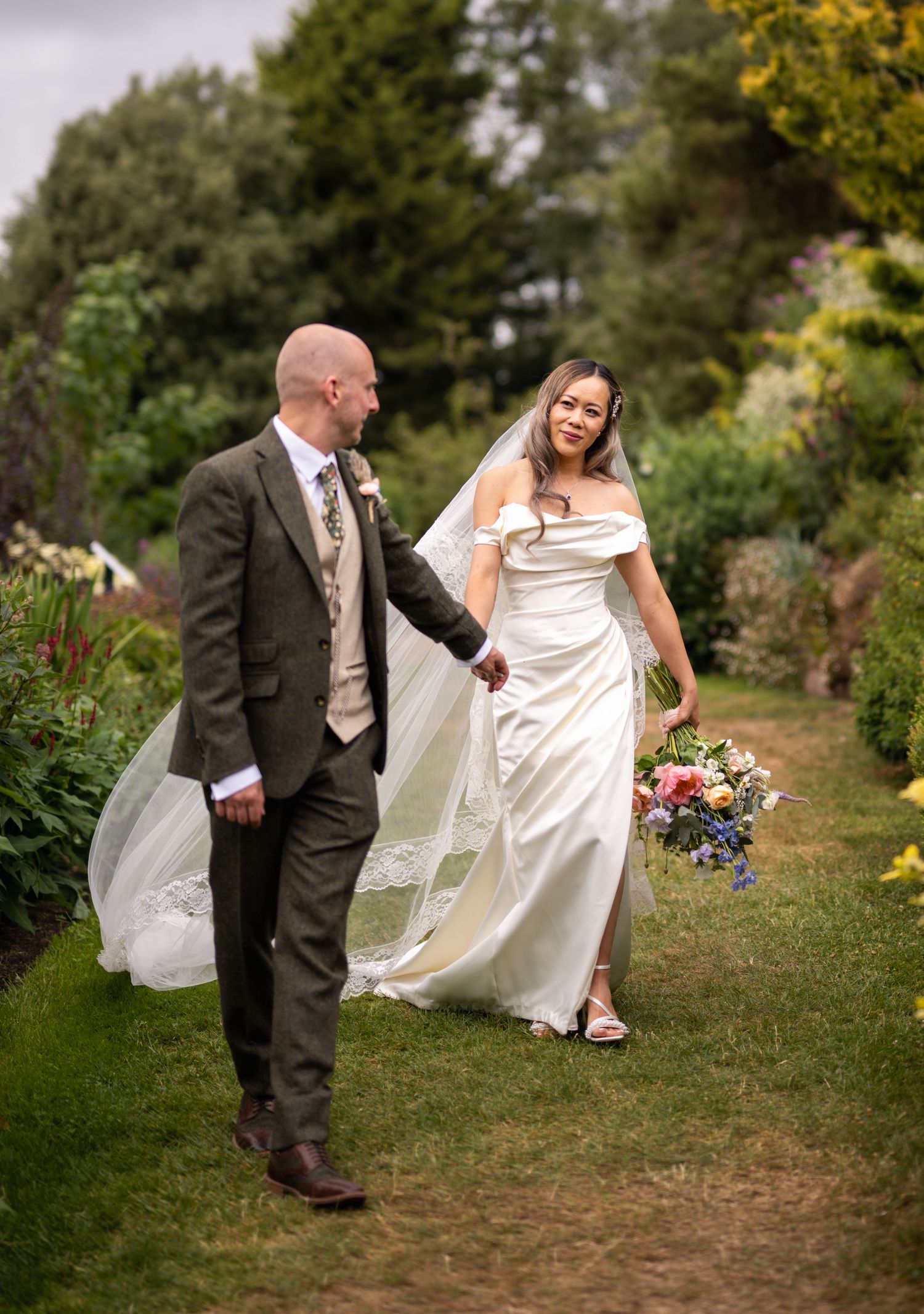 A bride and groom walk hand-in-hand through a lush garden, she in a white gown with veil, he in a grey suit.