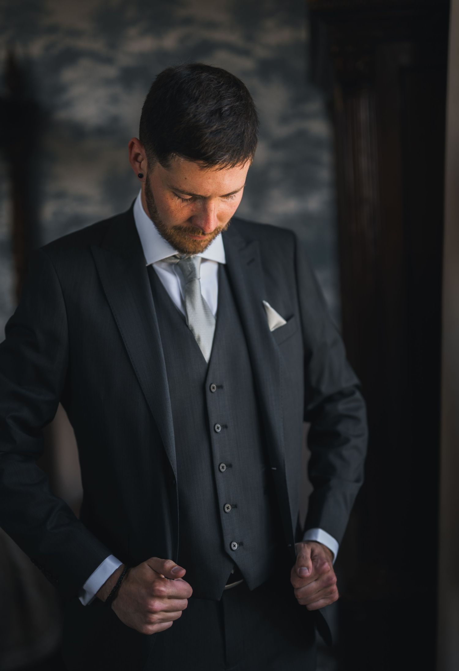 Dapper groom adjusting his dark three-piece suit with white pocket square before wedding ceremony.