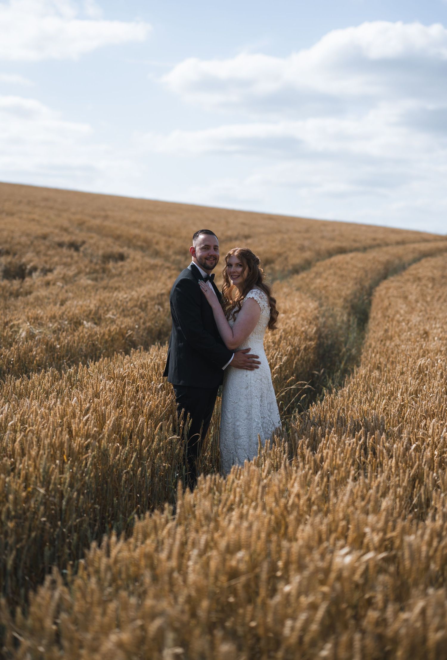 A bride and groom embrace in a golden wheat field under a cloudy sky on their wedding day.