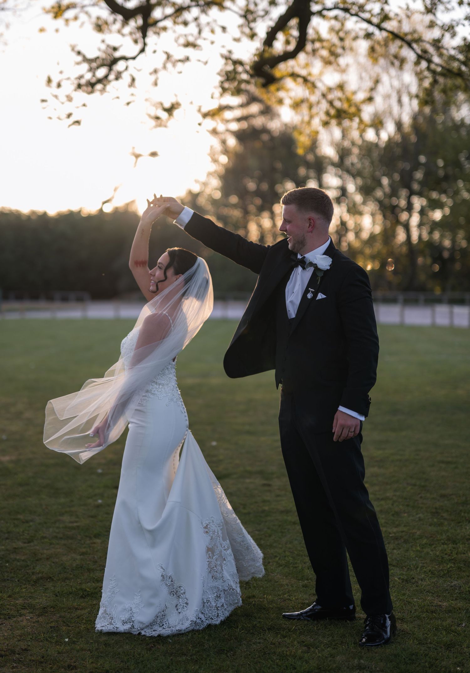 Bride and groom dancing outdoors at golden hour, groom twirling bride in white gown and veil on lush green lawn.