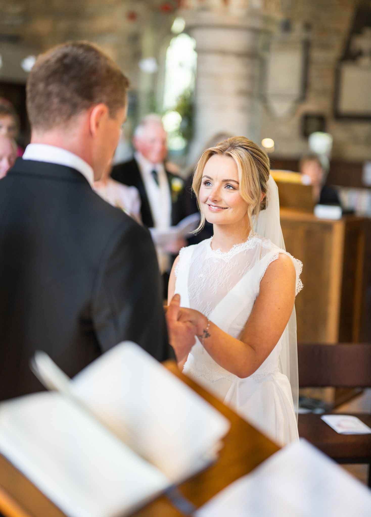 Bride smiling at groom during church wedding ceremony, wearing white dress and veil, holding hands at the altar.