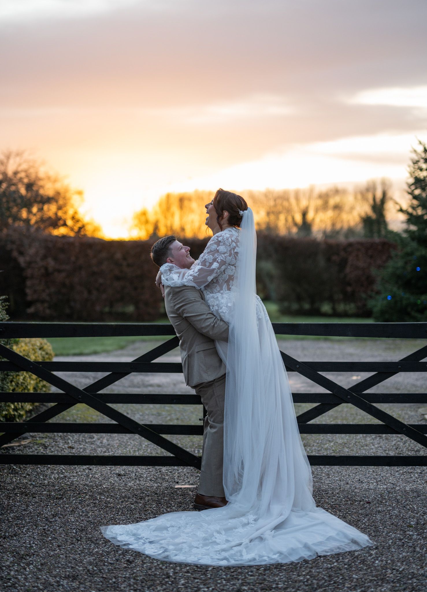 Groom lifting bride in white gown at sunset by garden gate, golden sky backdrop.