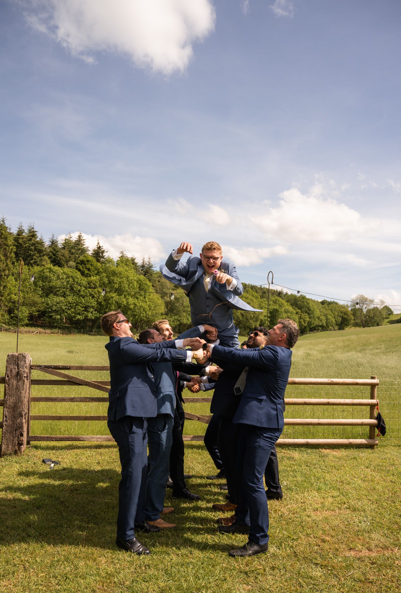 Groomsmen in blue suits tossing the groom into the air outdoors at a wedding celebration.