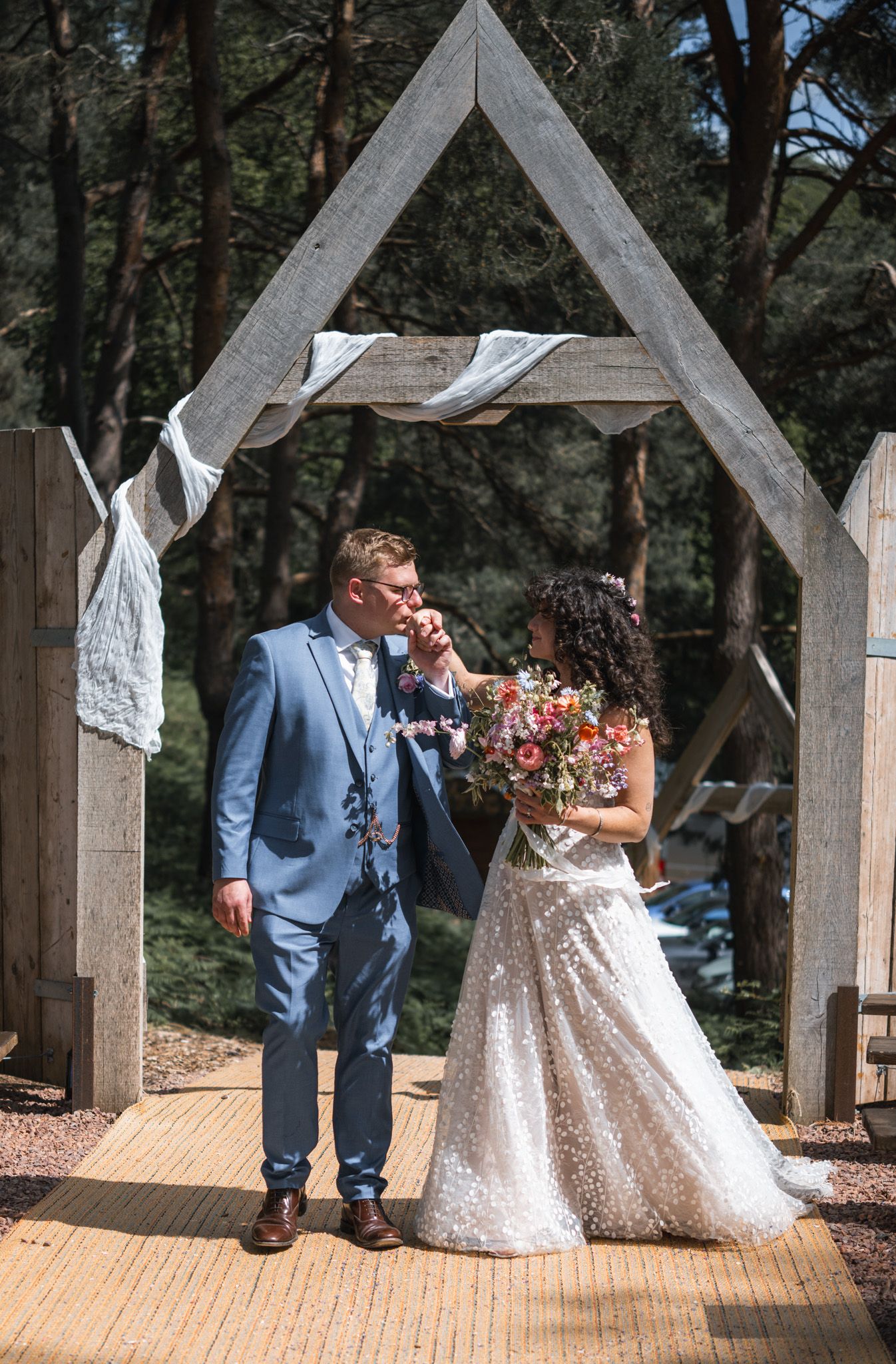 Bride and groom share a romantic moment under a wooden arch, bride holding a colorful bouquet, surrounded by trees.