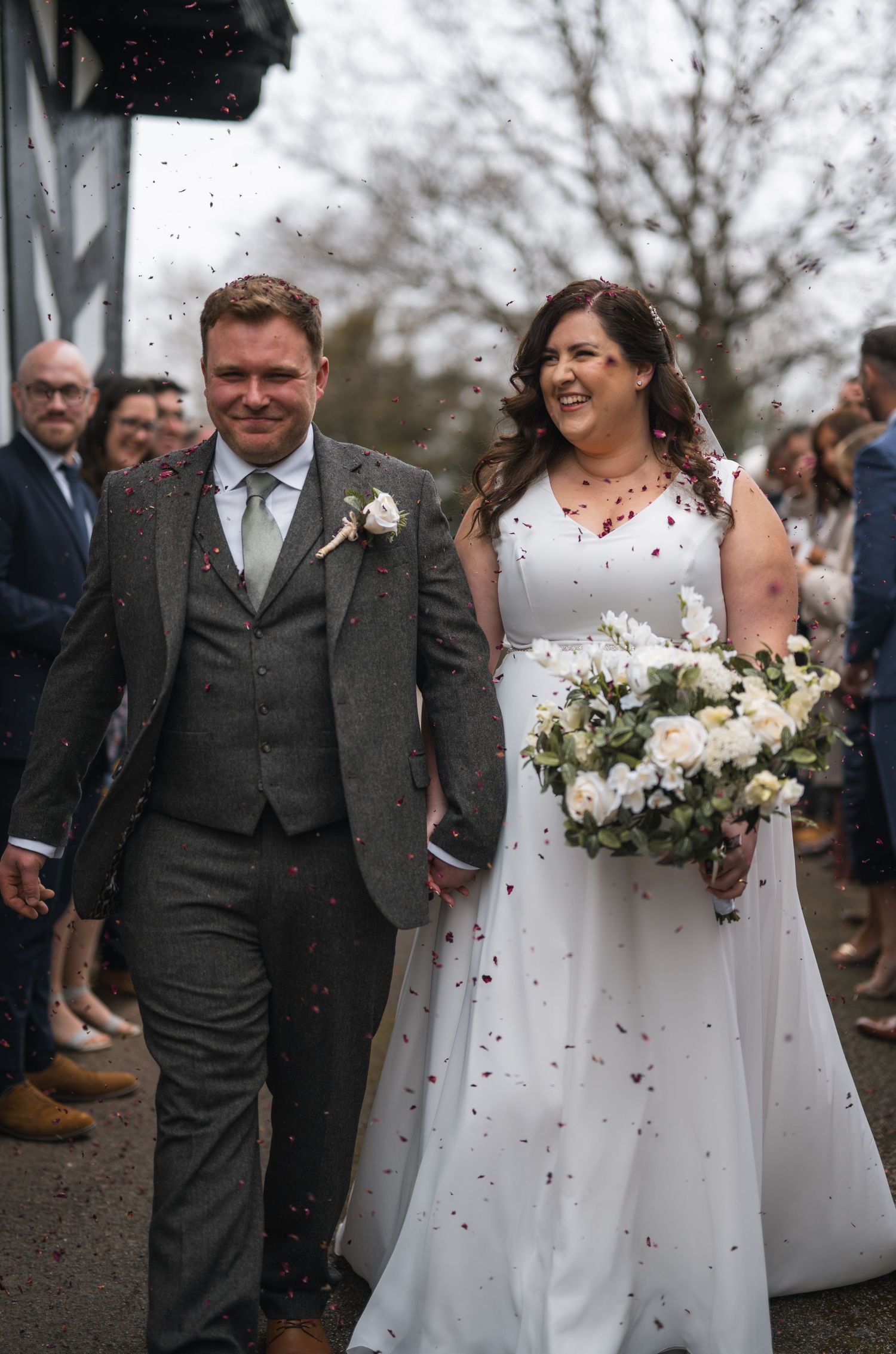 Smiling newlyweds walk through confetti shower, bride in white gown holding bouquet, groom in grey suit, guests cheering.