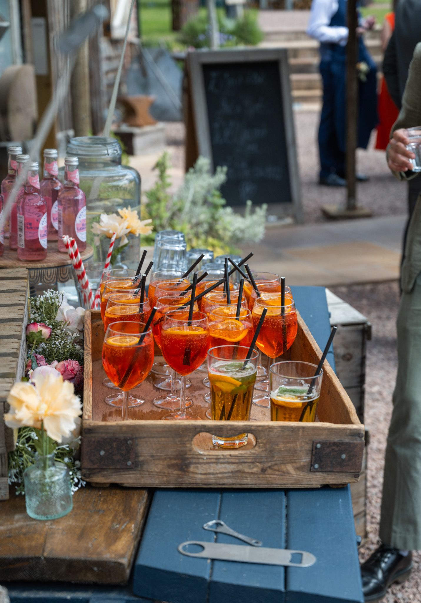 Rustic wooden tray with Aperol Spritz cocktails garnished with orange slices and black straws at an outdoor event.