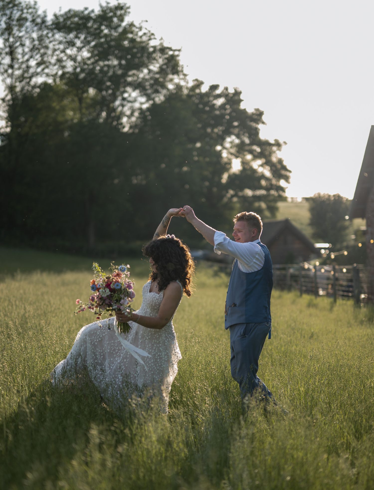 Bride and groom dancing in a sunlit meadow, she holds a wildflower bouquet in a lace gown as he twirls her.