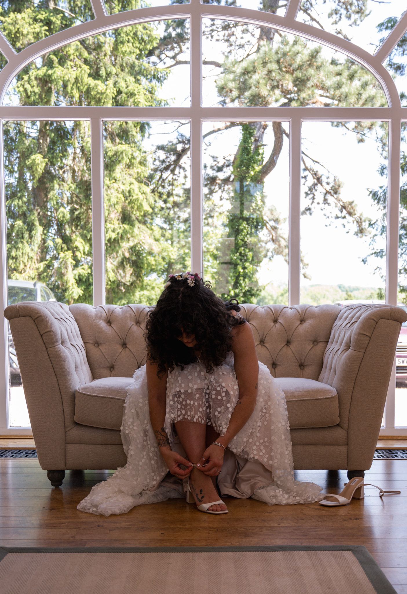 Bride in white lace gown adjusting heels on tufted sofa before large arched window with garden views.