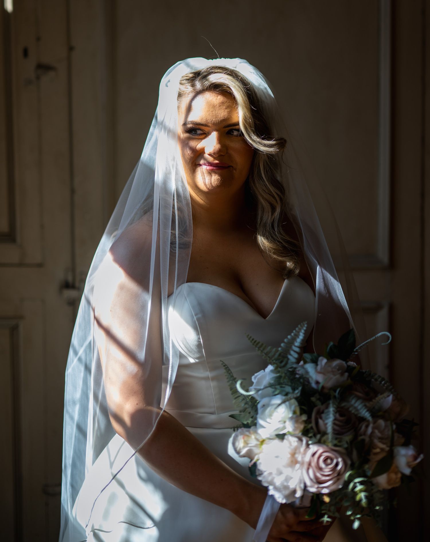 Bride in strapless white gown and veil holding pink and white bouquet in dramatic light.
