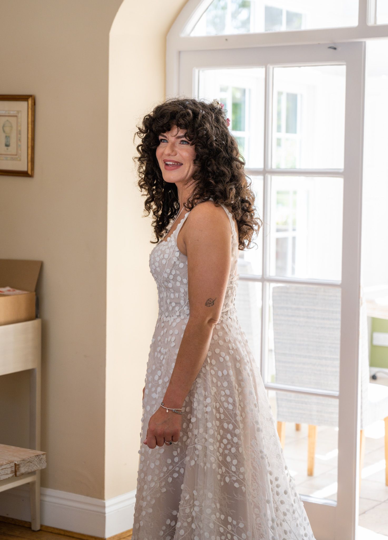 Woman in white floral dress with dark curly hair smiling near a bright window in an elegant room.