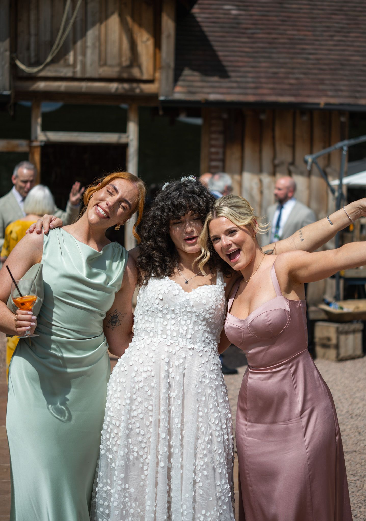 Three women in formal dresses smile and pose together outdoors at a wedding, with the bride in white floral gown.