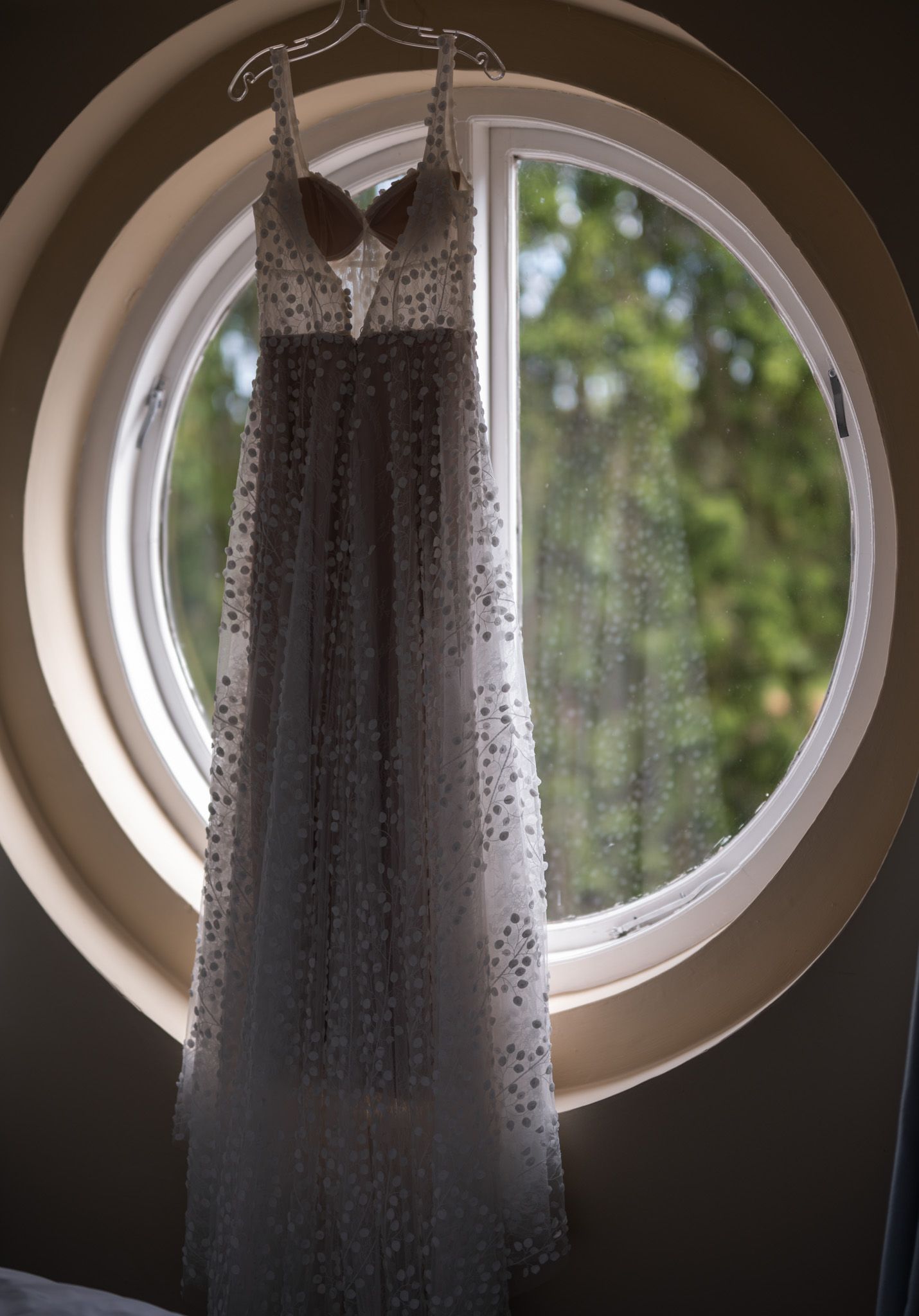 Elegant white dotted maxi dress hanging on a hanger in front of a round window with green trees outside.