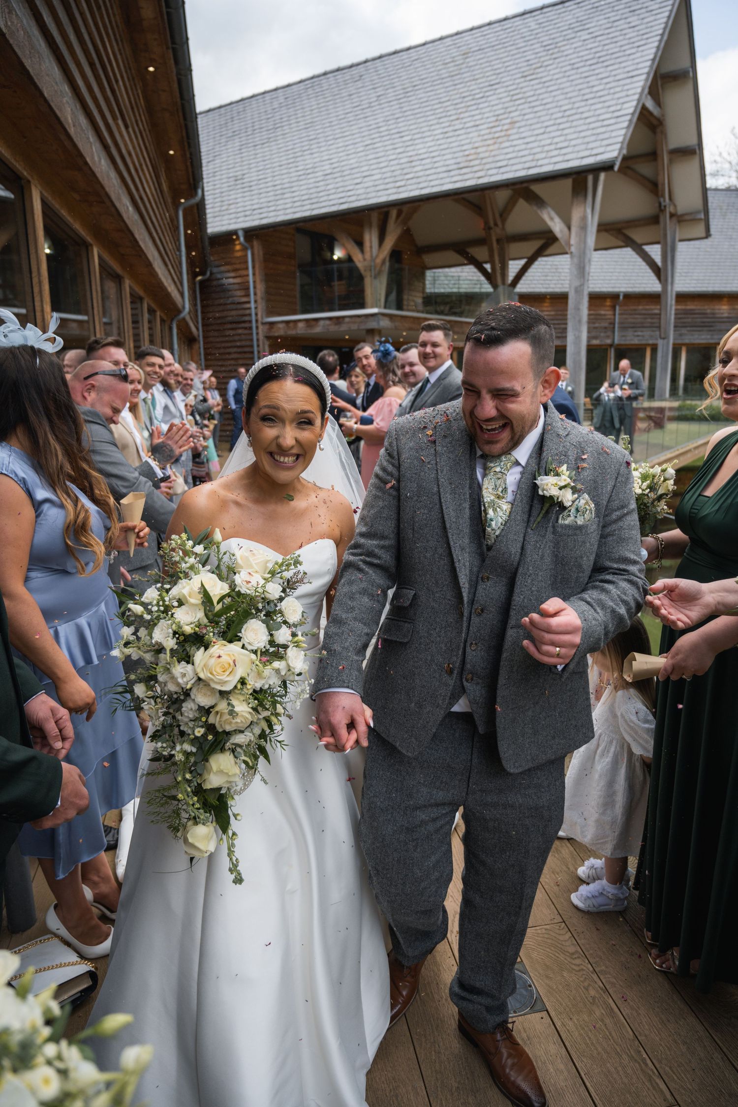 Joyful newlyweds walk through confetti shower outside rustic barn venue, bride holding white floral bouquet.