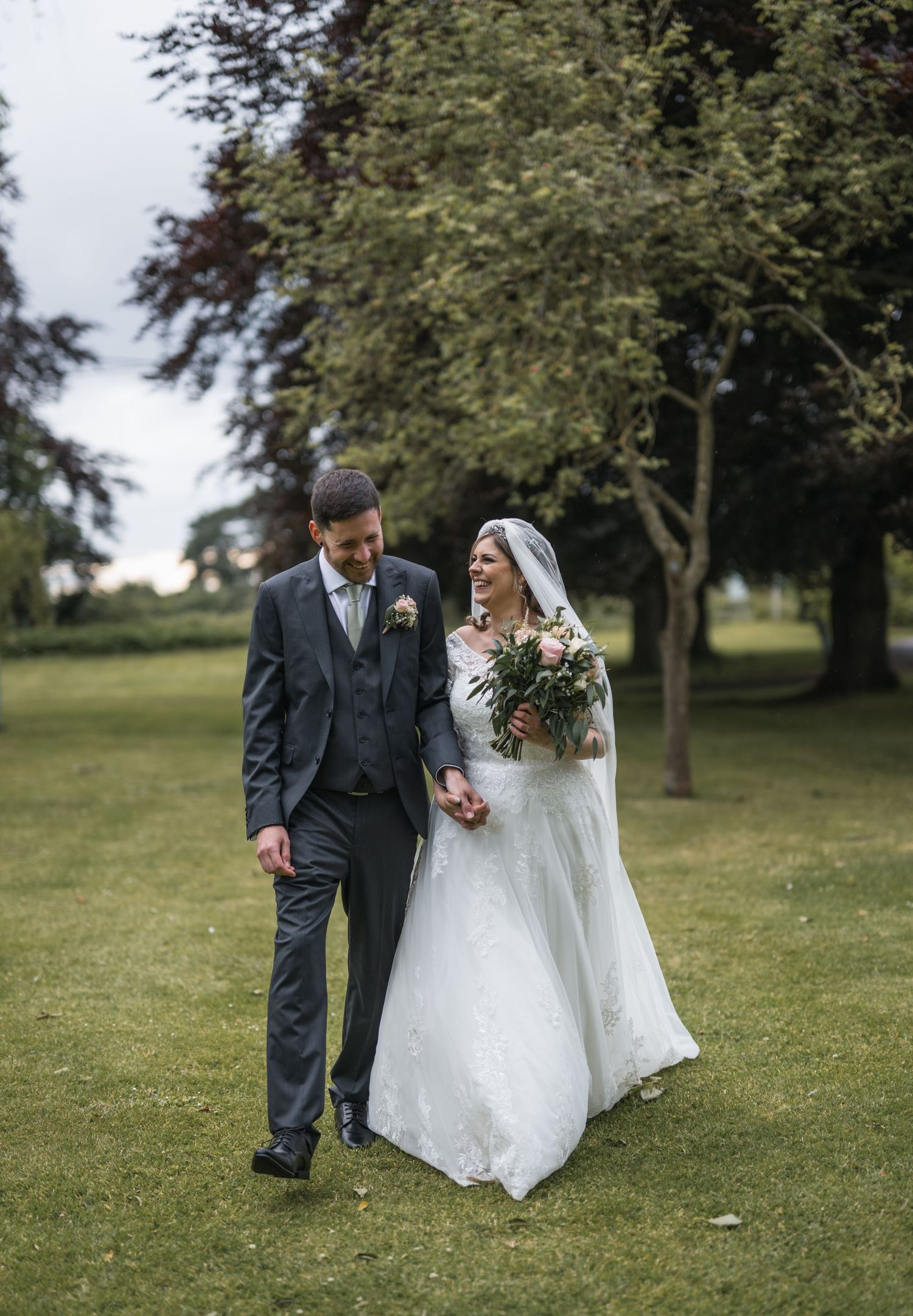 Bride and groom walk hand-in-hand through lush green gardens, bride holding bouquet in elegant white gown.