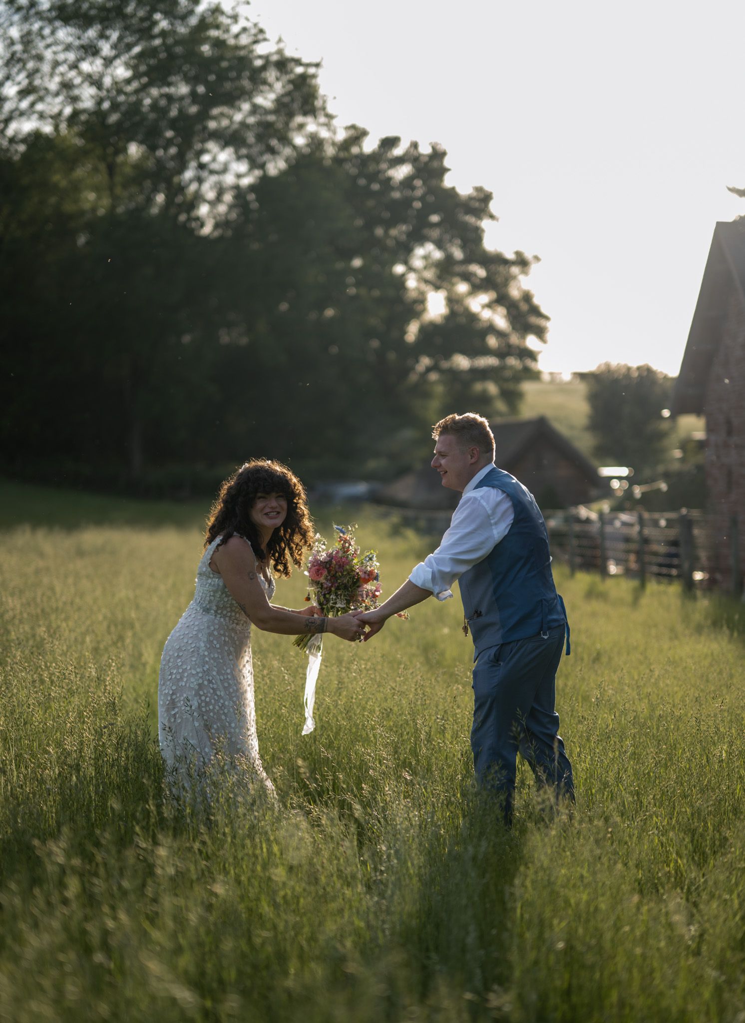 A bride and groom hold hands in a sunlit green meadow, the bride holding a colorful bouquet near a rustic barn.