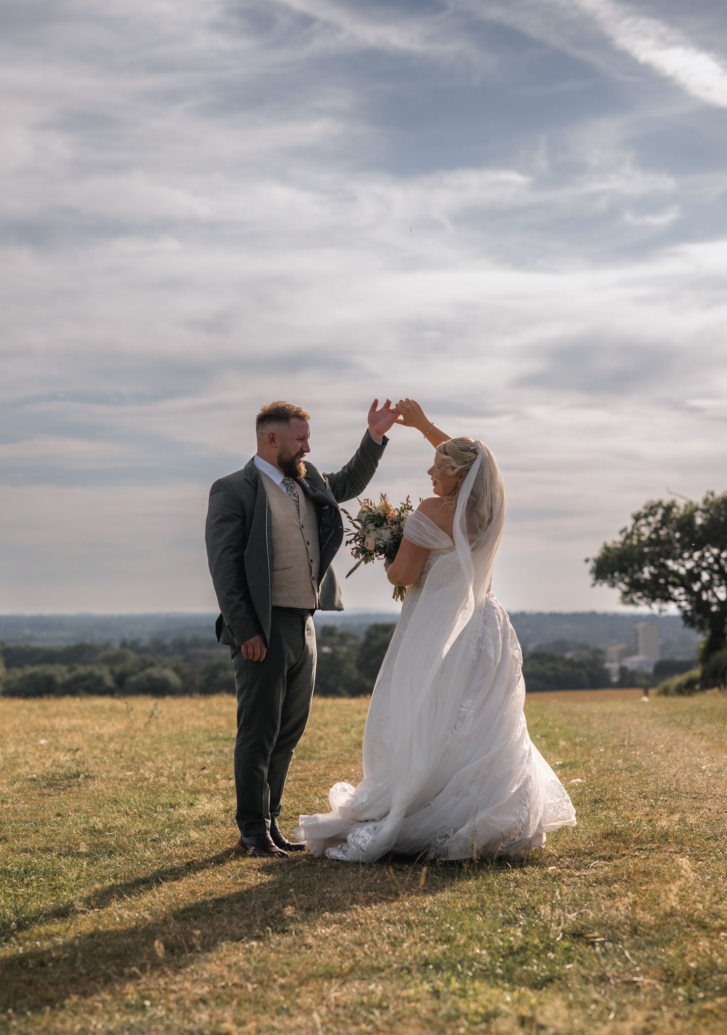 A bride in a white gown twirls as her groom holds her hand on a scenic hilltop during their outdoor wedding.