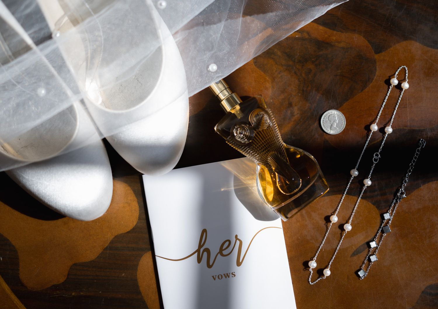 Flat lay of wedding details including white heels, veil, perfume bottle, coin, pearl necklace, and a her vows booklet on wood.