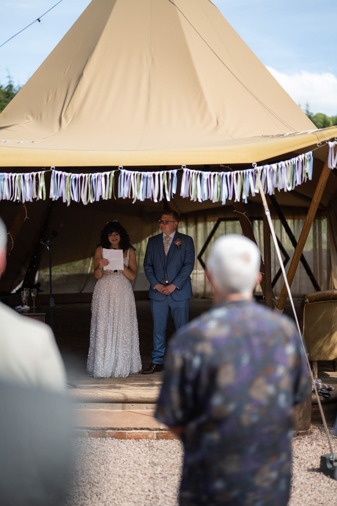 Couple stands under a decorated tipi tent during an outdoor wedding ceremony on a sunny day.